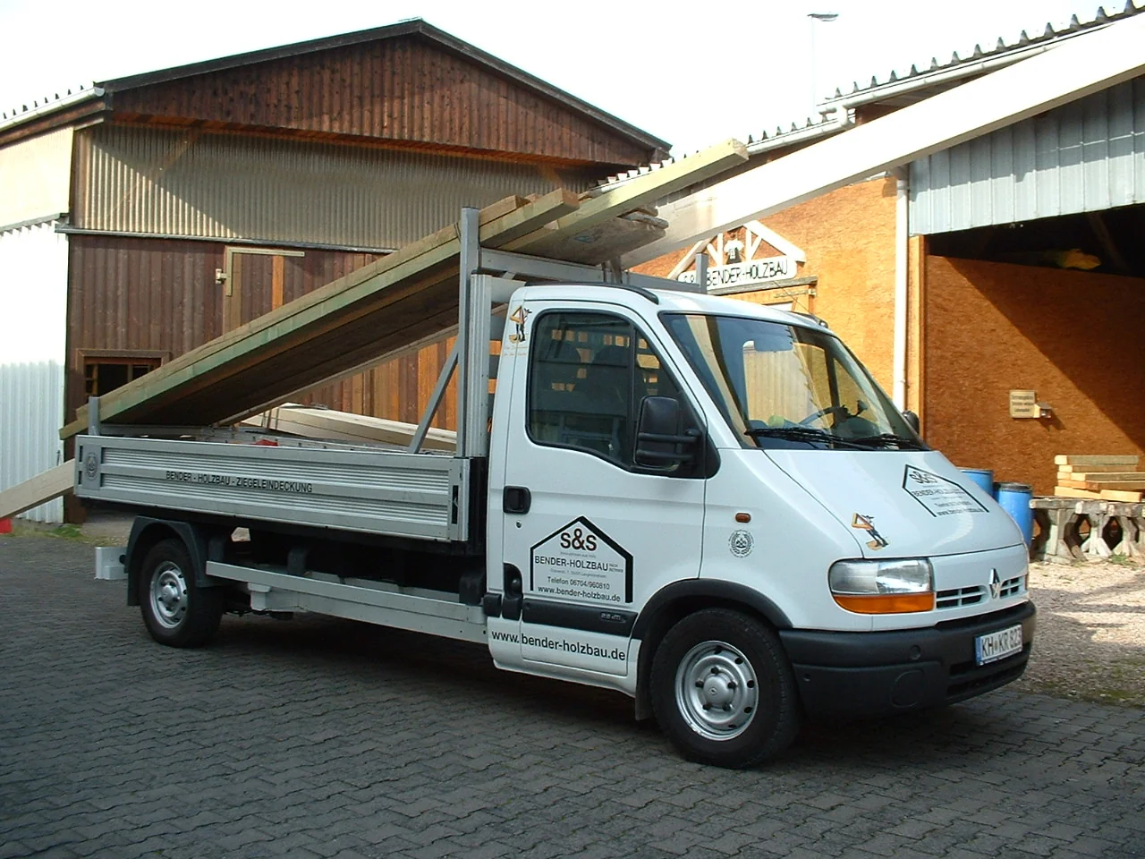 Pritschenwagen von S&S Bender-Holzbau transportiert lange Holzbalken vor einer Lagerhalle.