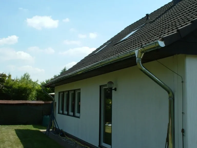Zinkfarbene Dachrinne mit Fallrohr an der Traufe eines Wohnhauses mit zwei Dachfenstern.