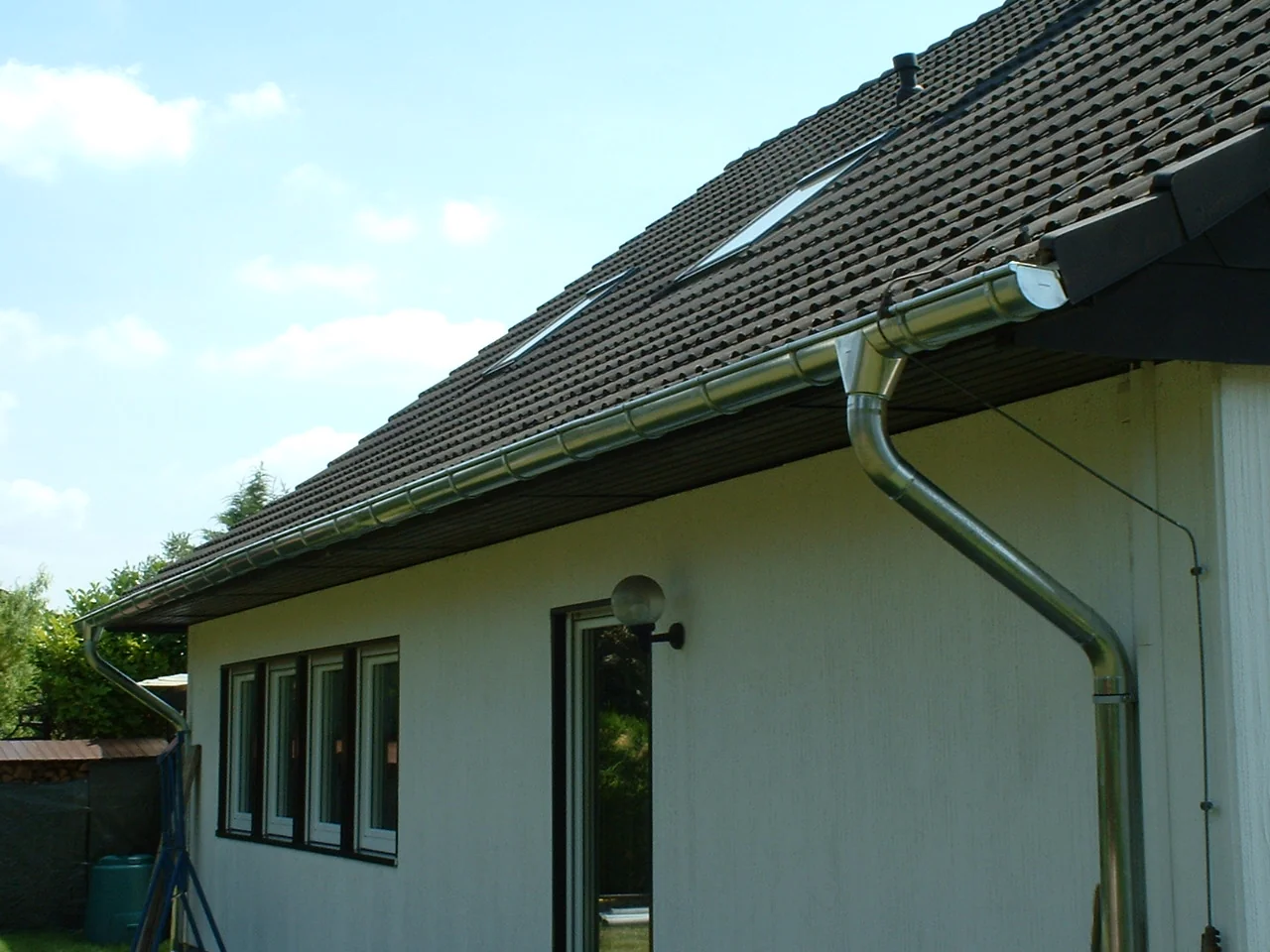 Neu montierte Zinkdachrinne mit Fallrohr an der Traufe eines weißen Wohnhauses.