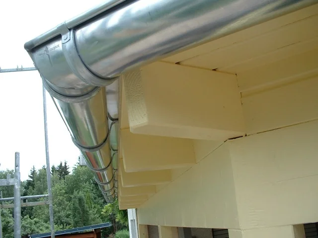 Neu montierte halbrunde Zinkdachrinne mit Fallrohr an der Traufe eines Hauses mit heller Holzverkleidung